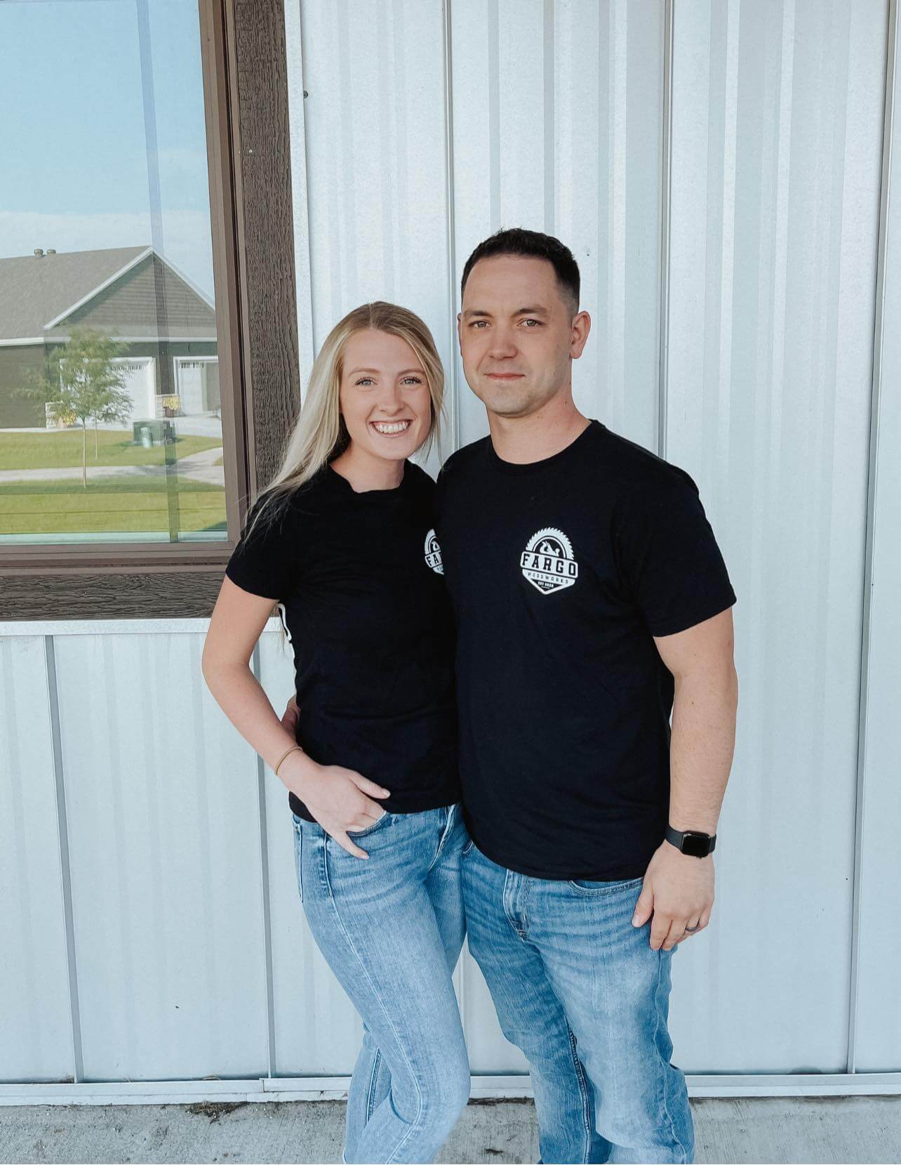 Jake & Melissa in front of a white corrugated metal wall with a window, evoking a sense of home. Both are wearing black T-shirts and blue jeans. The woman has long blonde hair, while the man sports short dark hair. They smile warmly, arms wrapped around each other.