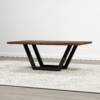 The Addison Coffee Table showcases a rectangular wooden surface with a substantial dark brown finish, complemented by a black metal V-shaped base. It is set against the backdrop of a light-colored speckled rug in an airy, minimalistic room featuring light gray flooring and a simple white wall.