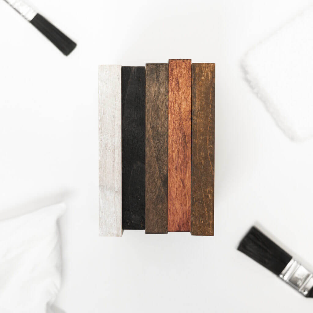A top-down view of six Wood/Finish Samples arranged vertically, each stained with a different color. These samples range from light gray to various shades of brown and black. Surrounding the planks are three paintbrushes and a white cloth, all placed on a white surface to emphasize the finish.