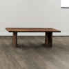 The Bennett Coffee Table, featuring a minimalist design with a rectangular top and two wide legs, stands on a wood-paneled floor against a plain wall. The table boasts a rich brown color with visible wood grain patterns, and the room is naturally lit from a nearby window.
