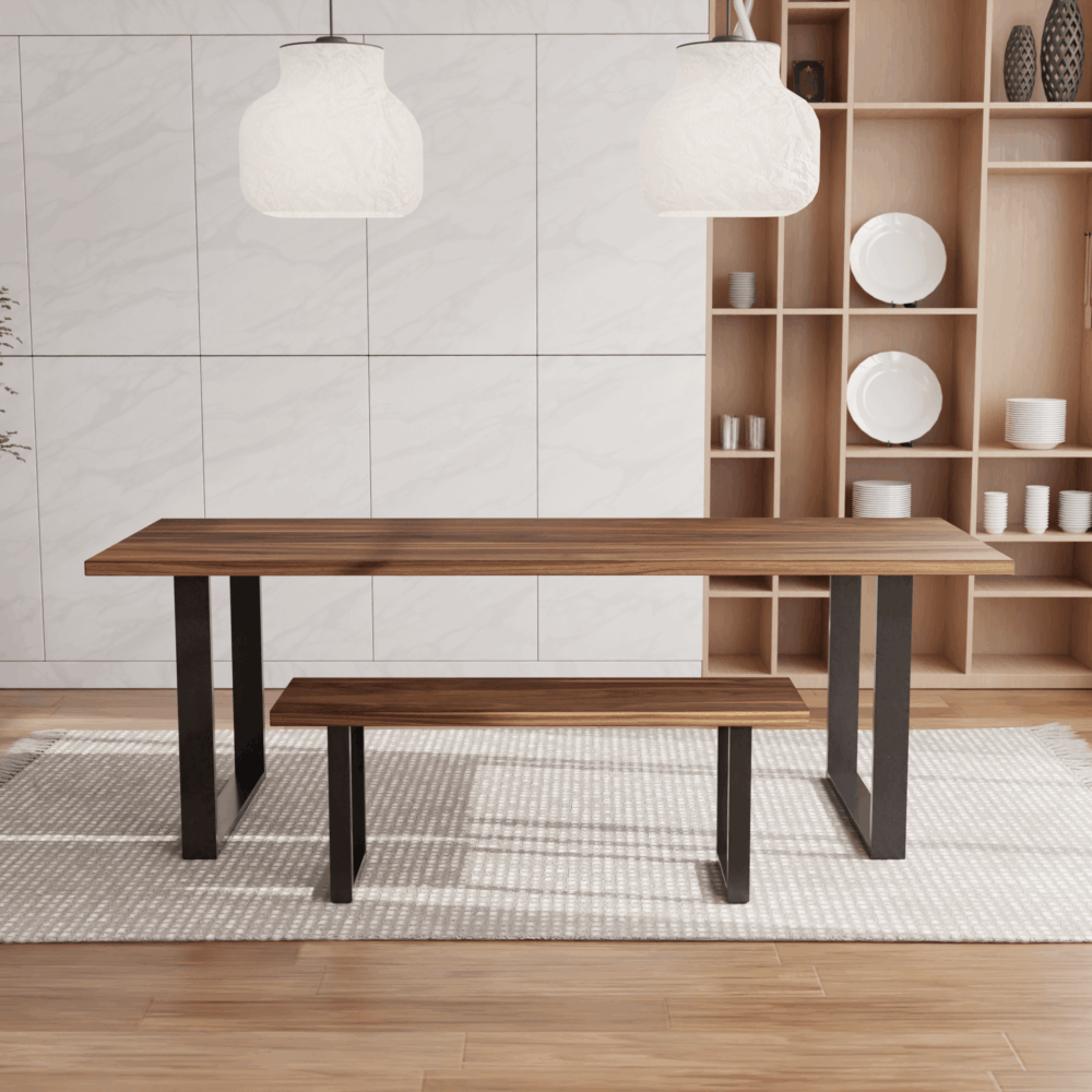 A contemporary dining area showcasing The Broadway Dining Table with black metal legs and a coordinated bench on one side. Two white pendant lights are suspended above. The backdrop features a light wood shelving unit adorned with decorative plates and cups, accompanied by a potted plant on the left.
