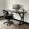 In a minimalist workspace with a modern desk, The Sienna Desk boasts sleek black metal legs and supports a monitor, keyboard, mouse, small notepad, and potted plant atop its surface. Positioned on a light wood floor against a plain white wall, the setup includes a black mesh office chair perfectly aligned in front of the desk.