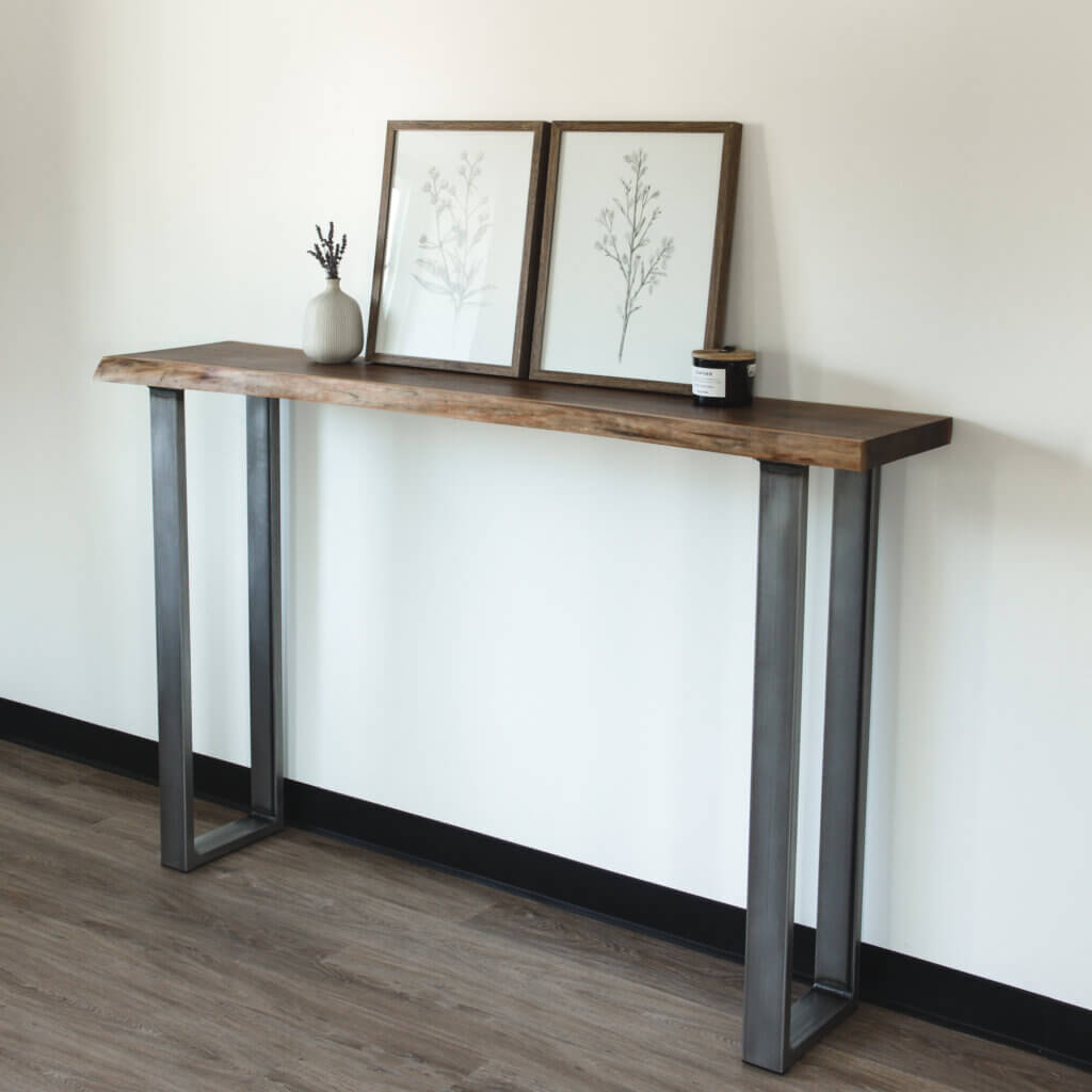 A minimalist Live Edge Walnut Sofa Table with metal legs stands against a plain white wall. Two framed botanical prints and a small vase with lavender sit on top of the table, alongside a small black candle. The floor features a light wooden texture.