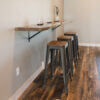 A minimalist kitchen setup features a Black Walnut Live Edge Floating Bar mounted to a wall with three matching wooden stools beneath it. The table holds a small plant and three glass candle holders. The hardwood flooring adds warmth to the modern, simplistic design.