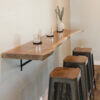 The **Black Walnut Live Edge Floating Bar**, mounted on a wall, showcases a rustic design paired with three metal and wood bar stools. The bar is adorned with three empty glass tumblers on coasters and a small white vase filled with greenery, creating an inviting and minimalistic ambiance.