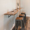 A minimalist seating area featuring a Black Walnut Live Edge Floating Bar attached to the wall, with three metal bar stools underneath. On the bar are three glasses and a small vase with greenery. The floor is wooden, and the walls are light beige.