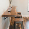 A side view of a modern kitchen counter featuring the Black Walnut Live Edge Floating Bar, paired with three rustic stools that have wood seats. Adorning the bar are a few decorative items, including cylindrical glass candle holders and a small plant in a vase. The floor is hardwood.