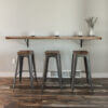 A minimalist setup featuring the Black Walnut Live Edge Floating Bar mounted on a light-colored wall. Three industrial-style metal stools sit beneath it. The bar holds four small glasses and a white vase with dried foliage, while the floor is made of rustic wood.
