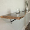 A Black Walnut Live Edge Floating Bar is elegantly mounted on a white wall using black metal brackets. This wooden shelf, crafted from rich black walnut and featuring a natural, live-edge finish, supports three glass candle holders with black bases that are evenly spaced along its length. In the background, a small plant vase can be seen on the far right end.