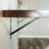 Close-up of the Black Walnut Live Edge Floating Bar mounted on a light-colored wall with black metal brackets. A glass cup is placed on the shelf, adding a touch of elegance. The background features a blurry view of a wooden floor and part of another wall.
