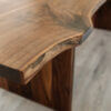 Close-up of The Live Edge Bennett Dining Table with its distinctive live edge design. The table’s surface reveals intricate natural wood grain patterns and variations in color, highlighting the craftsmanship and organic appearance of this exquisite furniture piece. The backdrop features a light-colored floor.