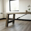 A modern, minimalist room featuring The Sheyenne Dining Table, known for its spacious rectangular wooden surface, sturdy legs, and striking diagonal metal support beam. The table is adorned with a decorative lantern and a small plant. A window in the background allows natural light to illuminate the space.