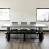 A minimalist dining area featuring The Sheyenne Dining Table flanked by black chairs and a matching bench on one side. The space is well-lit by natural light from two large windows on either side of the table, with light wood laminate flooring completing the aesthetic.