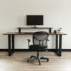A minimalist home office setup showcases The BIG Walnut Desk with black metal legs. On the desk are a monitor, a keyboard, a black mug, a small plant, and a closed book. In front of the desk is positioned a black mesh office chair. The room is complemented by light wood flooring.