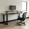 A minimalist workspace with The BIG Walnut Desk featuring black metal legs, a monitor on an elevated shelf, a keyboard and mouse on the main surface, a black office chair, an open notebook, a potted plant, and a black coffee mug. The room has a window and light wood flooring.