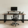 A modern, minimalist workspace featuring The BIG Walnut Desk with black metal legs, a single monitor on a riser, and a keyboard and mouse on top. An ergonomic mesh office chair is positioned in front of the desk. A small plant and a black mug add a touch of decor.