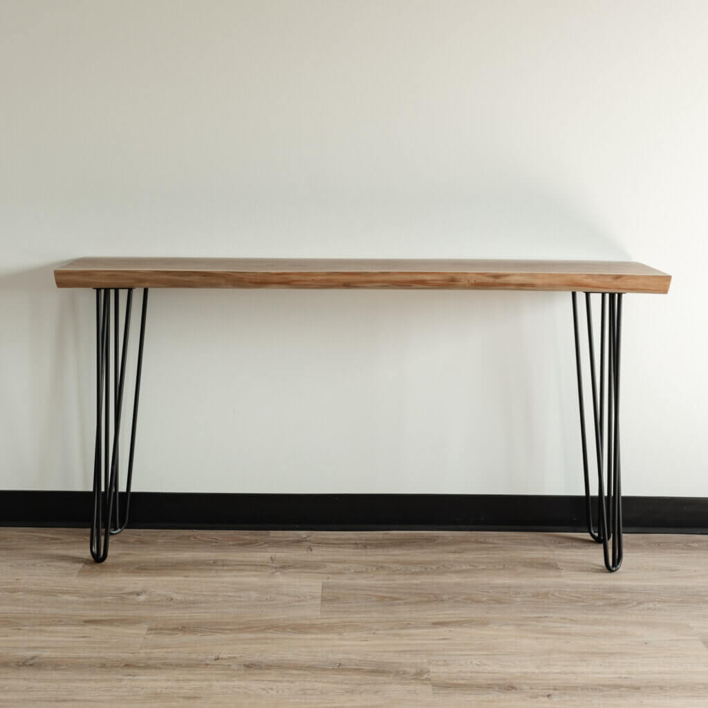 The Live Edge Black Walnut Hairpin Console Table, featuring a natural wooden top and sleek black metal hairpin legs, stands against a plain, light-colored wall on a wooden floor. The table showcases a clean, modern design with simple lines and a minimalist aesthetic.