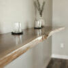 A Black Walnut Live Edge Floating Bar is mounted on a light gray wall. Two glass tumblers on coasters sit on the bar. In the background, a white vase with green foliage adds a decorative touch. The floor beneath appears to be wooden.
