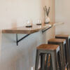 A Black Walnut Live Edge Floating Bar with metal brackets serves as a wall-mounted counter. On top of the bar, there are three candle holders and a vase with greenery. Below the bar, three brown wooden stools with metal legs are neatly aligned.