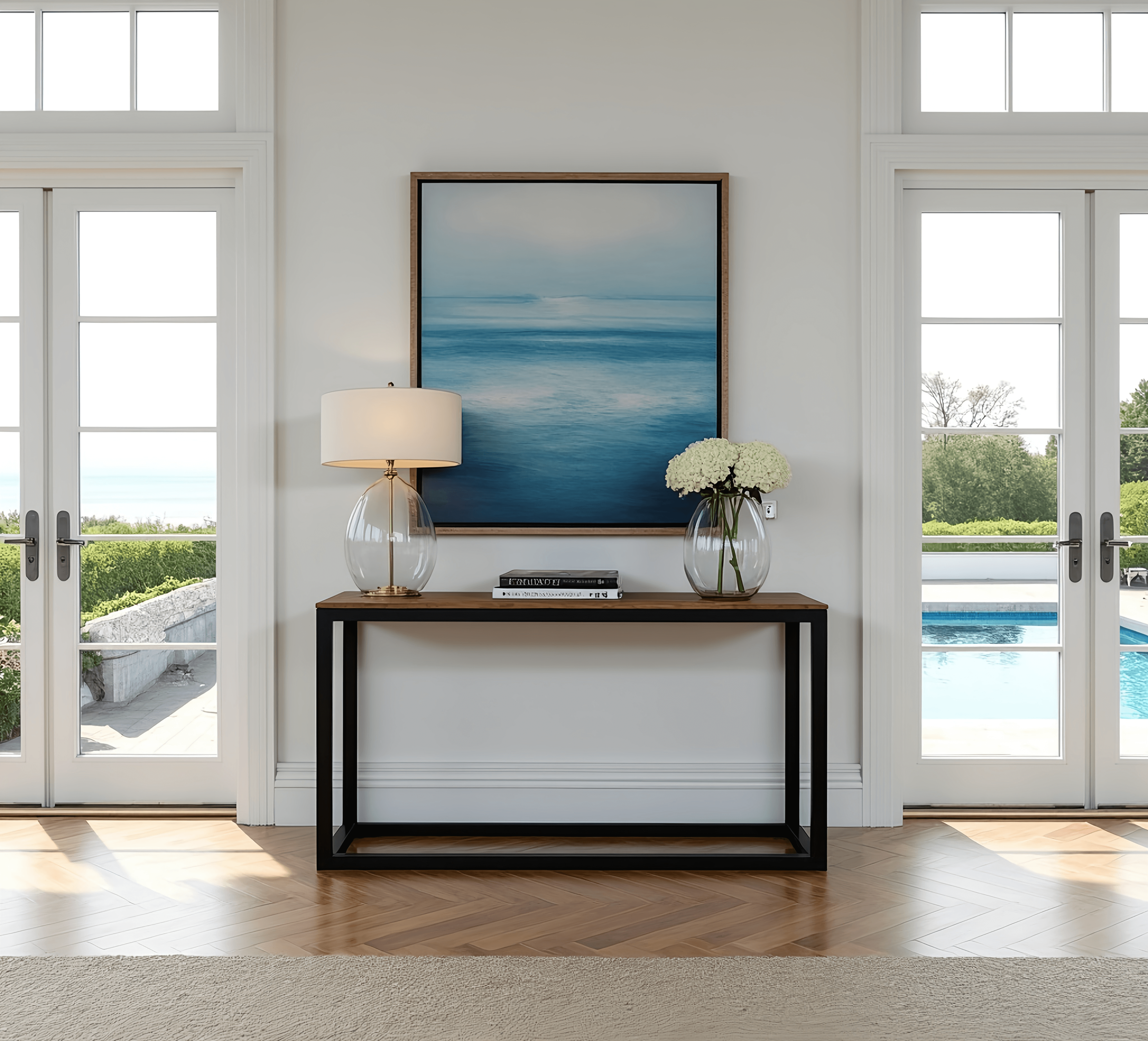 A contemporary room features The Sedona Console Table adorned with a lamp and a vase of flowers. A blue ocean-themed painting hangs above. Flanked by two glass doors, the scene opens to reveal a patio, pool, lush greenery, and a clear sky.