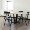 A minimalist dining room with The Monroe Dining Table and four black chairs. The room features light-colored walls, a large window, and hardwood flooring. The table has a black metal base, and the chairs have slender backrests and seats.
