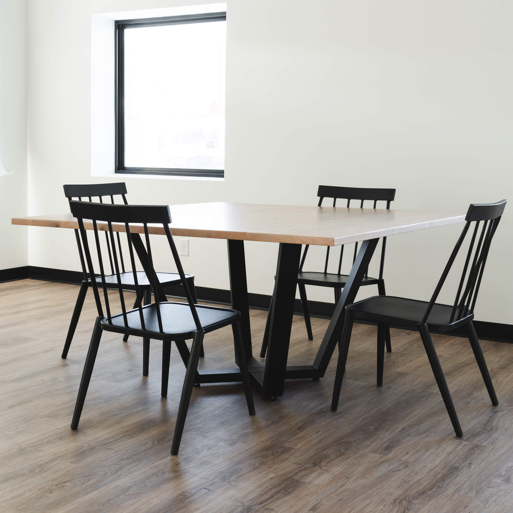 A minimalist dining room with The Monroe Dining Table and four black chairs. The room features light-colored walls, a large window, and hardwood flooring. The table has a black metal base, and the chairs have slender backrests and seats.