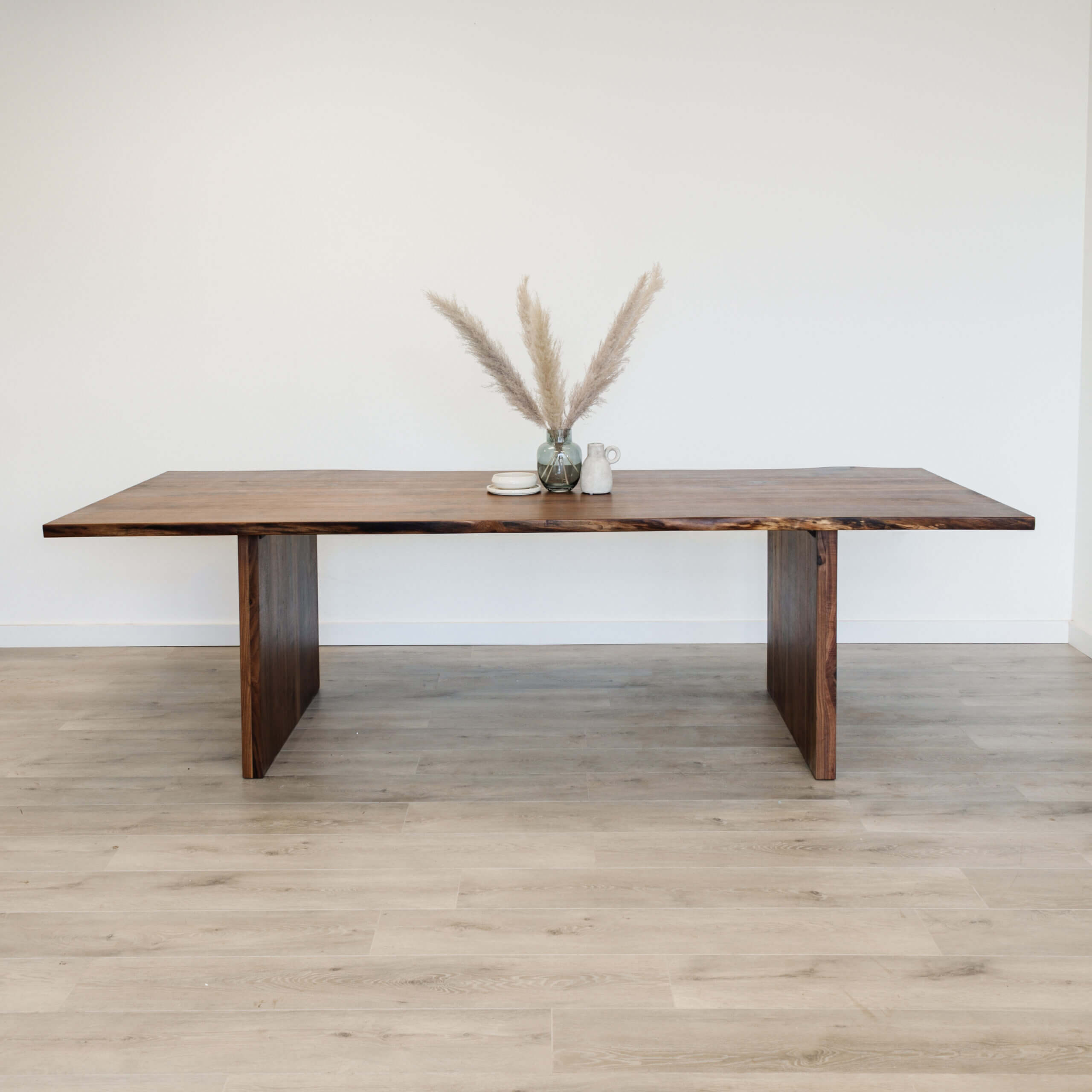 A minimalist scene features The Live Edge Bennett Dining Table at the center of a light-colored room with wooden flooring, showcasing a small arrangement of pampas grass in a vase on top.