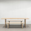 A minimalist wooden dining set, featuring The Benson Dining Table accompanied by two matching benches, all in a light natural finish. The set is showcased in a modern, open space with light gray walls and a wooden floor.