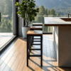 A modern kitchen is highlighted by The Sedona Bar Stools and large windows. A potted plant adorns the corner, offering a view of trees and mountains outside. Sunlight spills in, illuminating the hardwood floor.