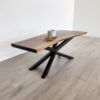 A modern The Dakota Live Edge Coffee Table with black metal X-shaped legs stands on a light wood floor. On the table is a white ceramic mug and a small beige rope decoration. The room has a minimalist and clean aesthetic with white walls.