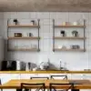 A modern kitchen with white tiled walls showcases an Industrial Hanging Pipe Shelf, complemented by two metal and wood open shelving units that hold potted plants, dishes, mugs, and glasses. Below the shelves lies a countertop featuring a microwave, kettle, and dish rack. Black-framed chairs are arranged around a wooden table.