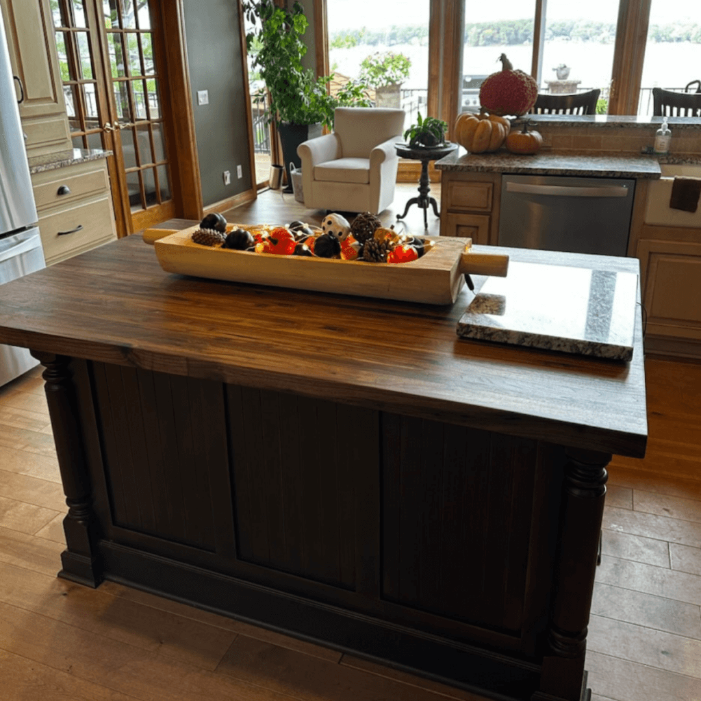A spacious, well-lit kitchen with a large central island featuring a dark wood countertop. The island is adorned with a wooden tray filled with decorative autumn items like pumpkins and pinecones. A white armchair and plenty of windows with a scenic view are in the background.
