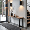 A modern interior features The Broadway Console Table against a white wall, accented by two vases with branches. Warm light from black wall sconces evokes Broadway charm. A large window reveals snow outside, while a staircase adds architectural interest nearby.