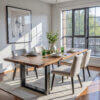 A contemporary dining room displays The Broadway Dining Table, characterized by its sleek black metal base. Surrounding it are cushioned chairs, accentuated by plates and green glasses. A vase of greenery highlights the center, with sunlight illuminating a light rug through expansive windows.