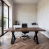Spacious modern office featuring The Modern X Desk and a black swivel chair on herringbone-patterned hardwood floors. A minimalist white sideboard with vases and a plant adorns the textured wall. Large windows provide a garden view.