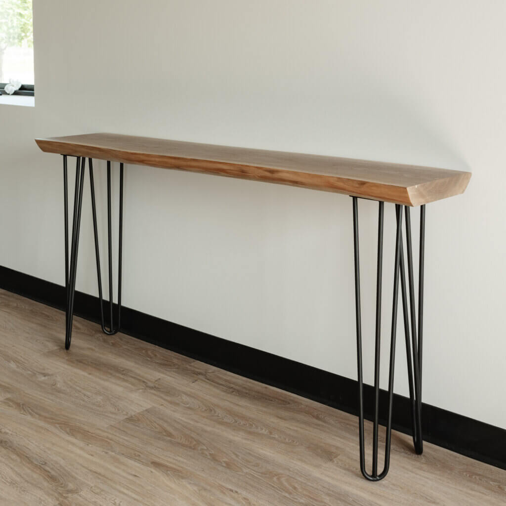 The Live Edge Black Walnut Hairpin Console Table, featuring a minimalist design with a natural finish and slim black metal hairpin legs, sits against a plain white wall on a light wood floor. The design is sleek and modern.