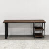 The Loredo Desk is a sleek, wooden office desk featuring contrasting black metal legs and two wooden shelves on the right side. Positioned against a plain white wall, it pairs perfectly with the patterned area rug placed in front.