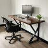 The minimalist workspace includes The Sienna Desk with a wooden top and black metal legs, paired with a computer monitor, keyboard, mouse, and small plant. It also features a black ergonomic office chair on a polished wooden floor.