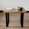 The White Oak U-shaped End Table with its small white oak tabletop and U-shaped black metal legs stands on a wooden floor. Atop the table are a notebook with a pen, a lit candle in a jar, and a black coffee mug resting on its saucer. The background showcases a plain white wall.