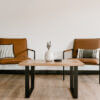 A minimalist sitting area showcasing two brown leather chairs with striped cushions, positioned on either side of The Broadway Coffee Table, featuring chic black metal legs. A white vase with dried flowers and a small black object adorn the table, creating an elegant Broadway-inspired setting.