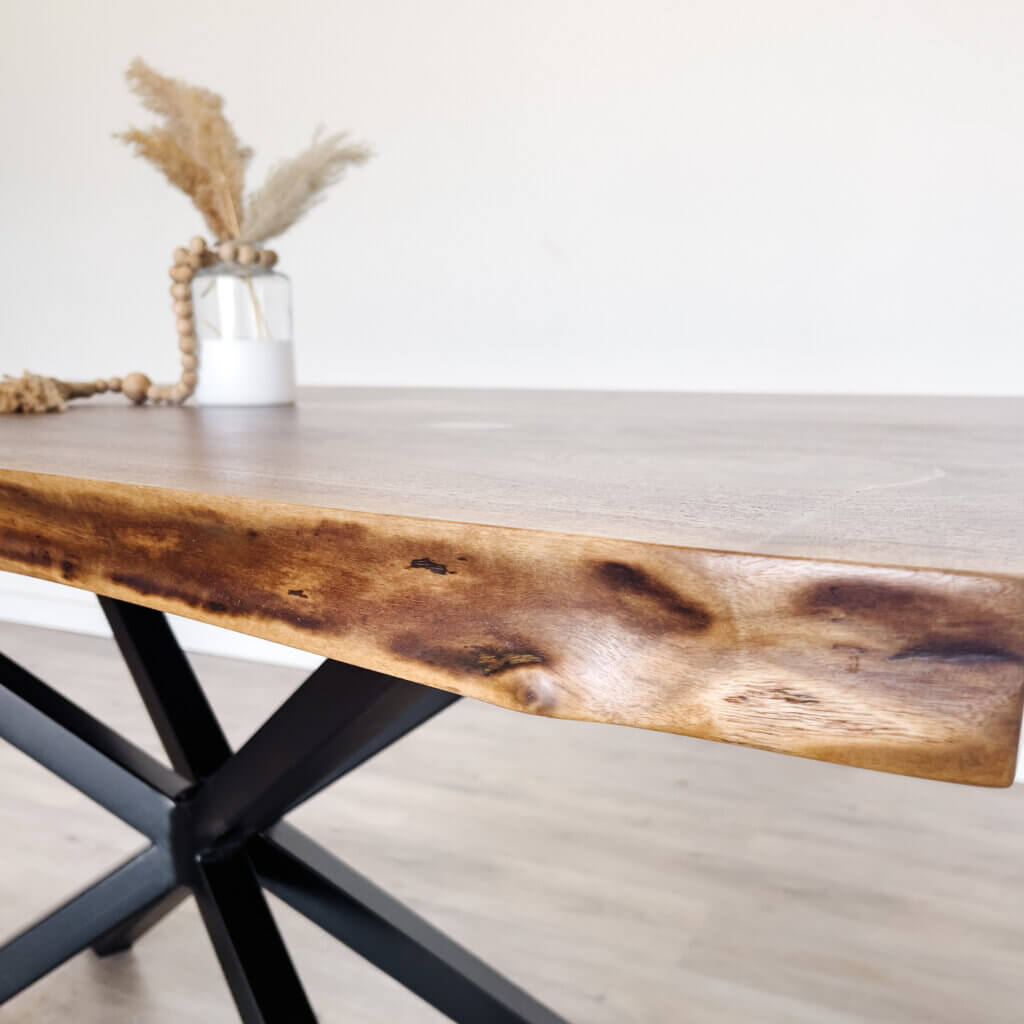 Close-up of a wooden dining table with a natural, irregular edge, showcasing the latest trends in live edge furniture. The tabletop has light wood grain patterns, and the black metal legs form an X-shape. In the background, a small white vase containing dried grasses and a string of wooden beads is placed on the table.