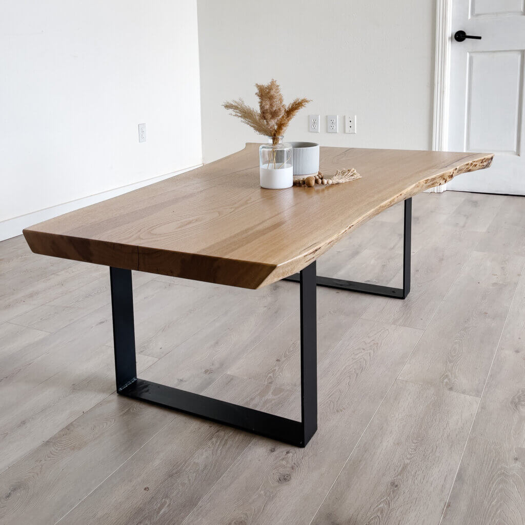 A rectangular wooden coffee table with natural live edges on the top, resting on black metal legs. On the table sits a white candle and a small beige vase containing dried pampas grass and wooden beads. The table is placed on a light wood floor in a minimalist room, embodying current trends.