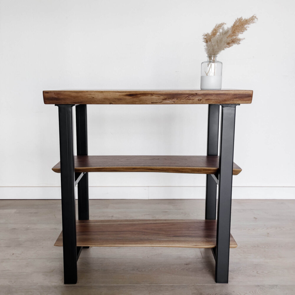 A three-tiered wooden shelf with black metal legs stands against a plain white wall on a light wood floor. A glass vase with dried pampas grass decorates the top shelf, adding a minimalist touch to the modern furniture piece, perfectly aligning with current live edge furniture trends.