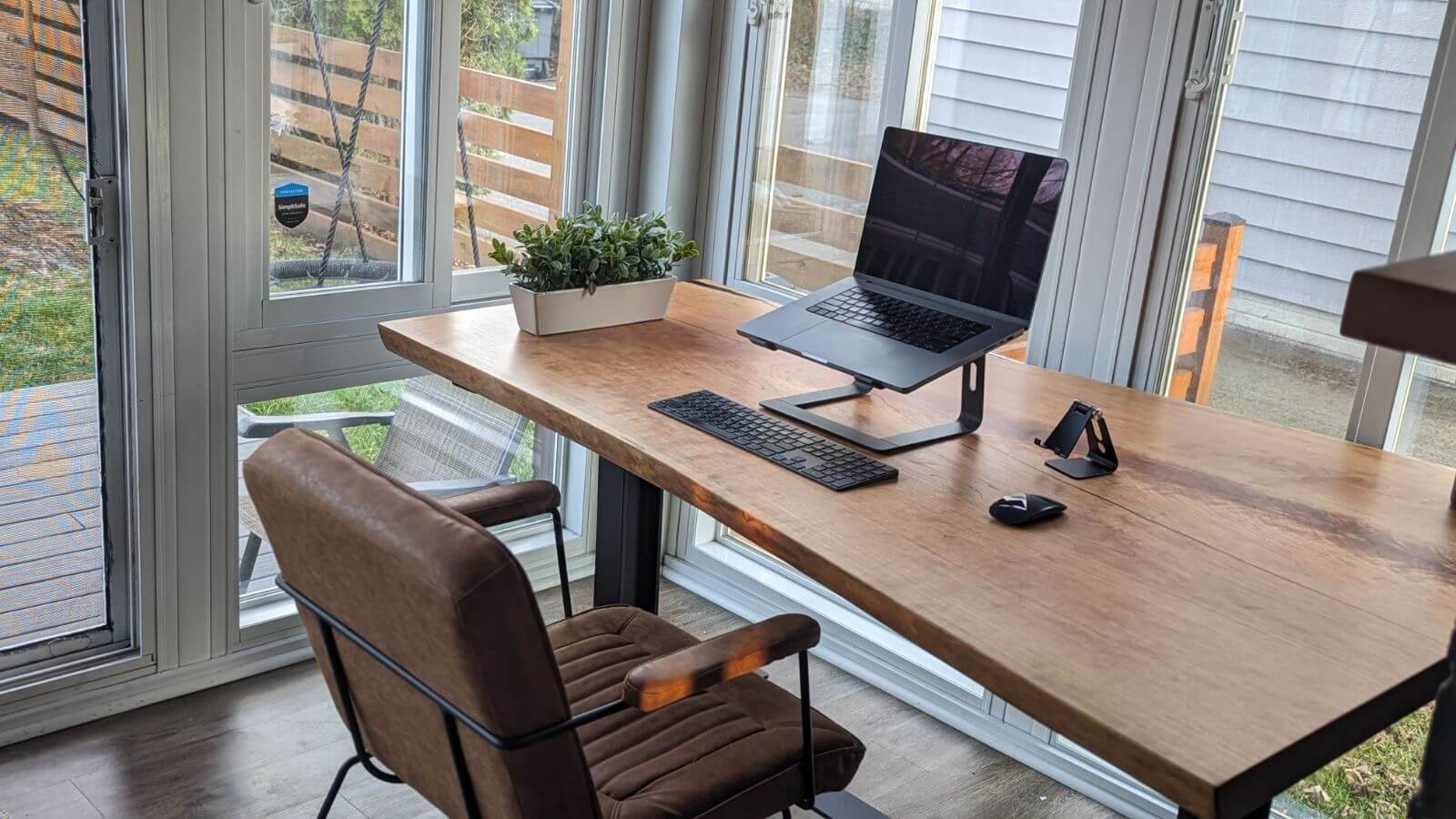 A neatly arranged home office setup by a large window features the latest in furniture trends. The desk holds a laptop on a stand, a keyboard, a mouse, and a small potted plant, all atop an elegant piece of live edge furniture. A brown cushioned chair is placed in front of the desk. Natural light floods the room from the surrounding windows.