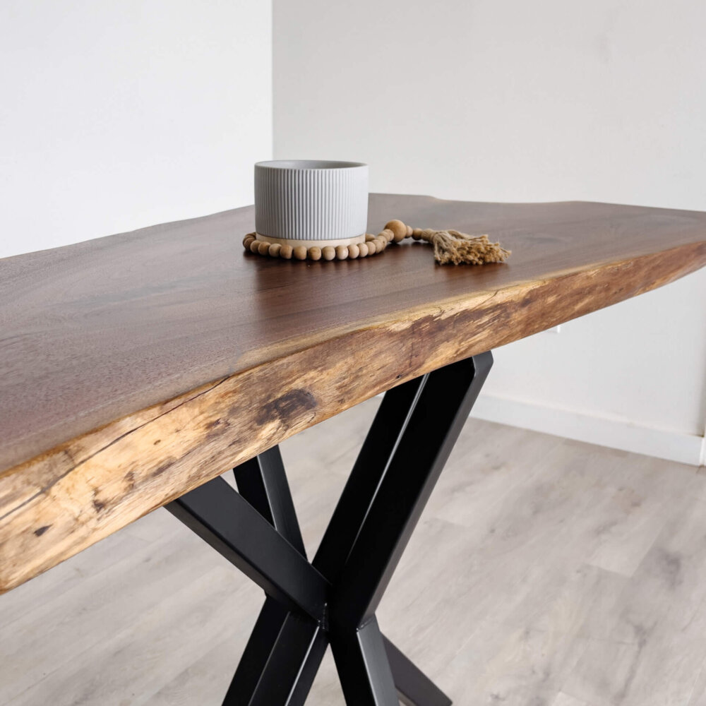 A modern wooden table with a live edge, held by black metal legs, stands on a light wood floor. A small gray ceramic pot and a wooden bead garland with a tassel are placed atop the table. Embracing current furniture trends, this piece shines against the plain white wall in the background.