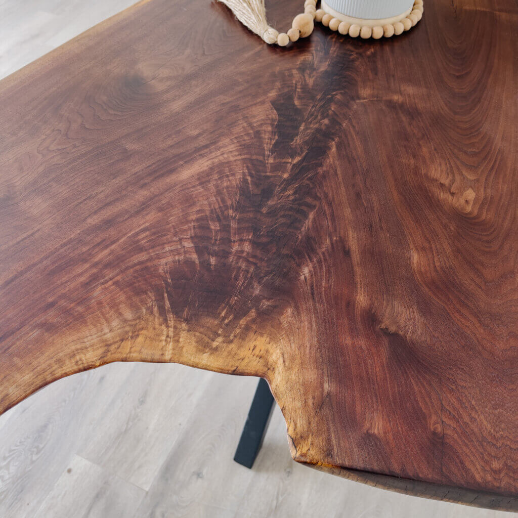 Close-up of a wooden table featuring rich, natural grain patterns and an irregular live edge, embodying the latest furniture trends. The surface has a warm, polished finish. A decorative piece with small wooden beads and part of a white vase is partially visible on the table.