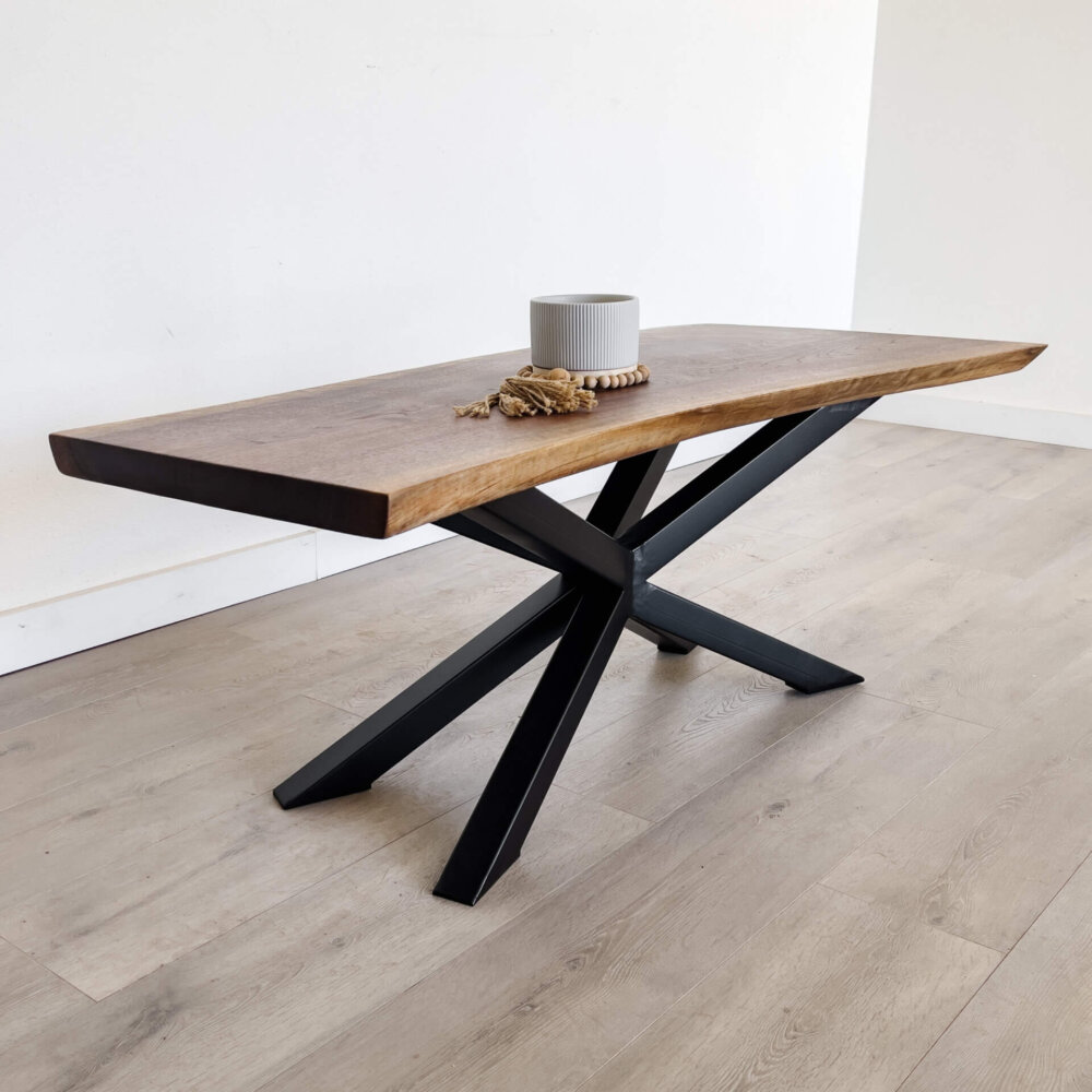 A stunning piece of live edge furniture, this wooden table boasts a natural edge top and a black, geometric X-shaped metal base. Positioned on a light-colored wooden floor, it showcases a gray ribbed cylindrical candle, a beaded decoration, and a small bundle of dried wheat.
