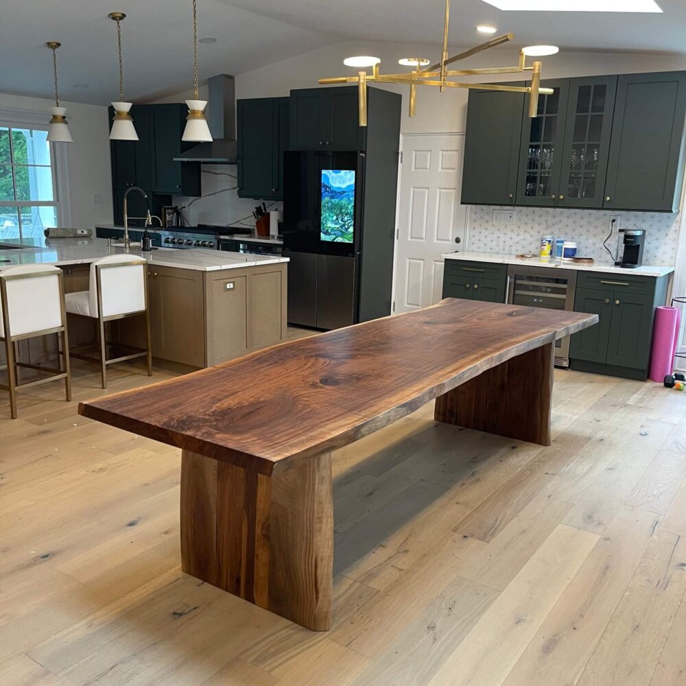 A modern kitchen with green cabinetry, light wood flooring, and a large wooden dining table showcasing the latest furniture trends with its natural live edge. White upholstered chairs are at the kitchen island, and contemporary pendant and chandelier lights are above the island and table.