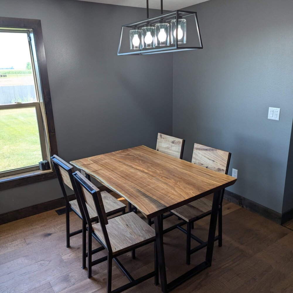 A modern dining room with a rectangular live edge wooden table and four matching chairs. The room has gray walls, a wooden floor, a window with a view of green grass outside, and an elegant rectangular light fixture hanging from the ceiling.