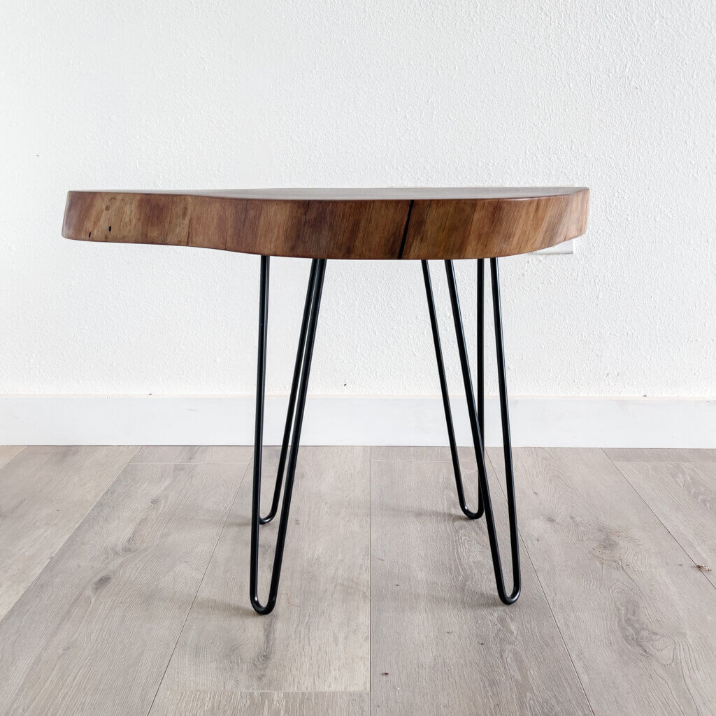 A small wooden side table with a natural, live-edge top and three black metal hairpin legs stands on a light wood floor against a plain white wall. This piece of Live Edge Furniture perfectly captures minimalist and modern trends.